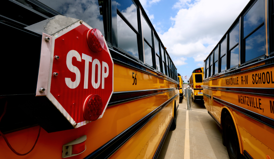 A close-up of a school bus stop sign with other yellow school buses parked in a row under a cloudy sky.